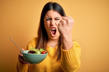 Young beautiful woman eating healthy fresh salad over yellow background annoyed and frustrated shouting with anger, crazy and yelling with raised hand, anger concept