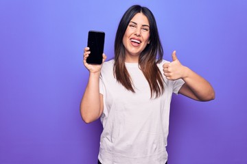 Young beautiful brunette woman holding smartphone showing screen over purple background smiling happy and positive, thumb up doing excellent and approval sign