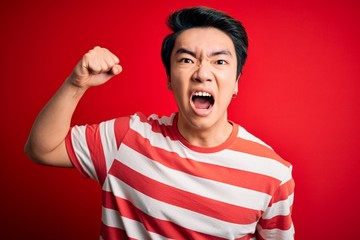 Young handsome chinese man wearing casual striped t-shirt standing over red background angry and mad raising fist frustrated and furious while shouting with anger. Rage and aggressive concept.