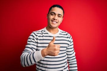 Young brazilian man wearing casual striped t-shirt standing over isolated red background doing happy thumbs up gesture with hand. Approving expression looking at the camera showing success.