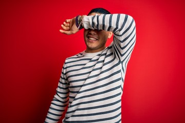 Young brazilian man wearing casual striped t-shirt standing over isolated red background covering eyes with arm smiling cheerful and funny. Blind concept.