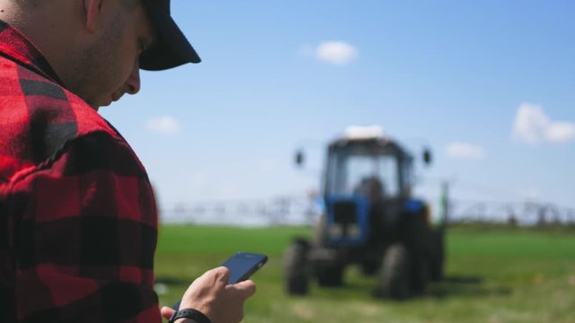 Young attractive farmer with phone standing in field, tractor working in green field in background. Smart farming using modern technologies in agriculture. Seasonal agricultural works.
