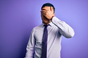 Young brazilian businessman wearing elegant tie standing over isolated purple background covering eyes with hand, looking serious and sad. Sightless, hiding and rejection concept