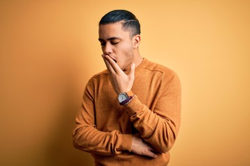 Young brazilian man wearing casual sweater standing over isolated yellow background bored yawning tired covering mouth with hand. Restless and sleepiness.