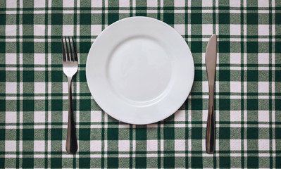 A white plate, knife and fork lie on a checkered green cotton tablecloth. Empty dish for copy space and mockup, top view.