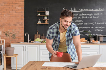 Young man with cup of hot coffee and laptop in kitchen