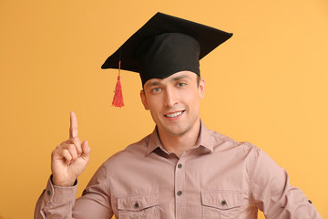 Young man in graduation hat and with raised index finger on color background