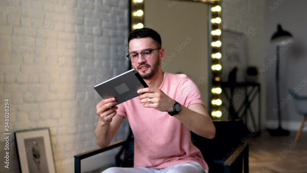 Poster positive young man in eyeglasses laughing while relaxing on chair in modern apartment and holding to