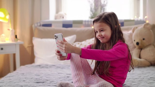Portrait of small girl with smartphone sitting on bed, taking selfie.