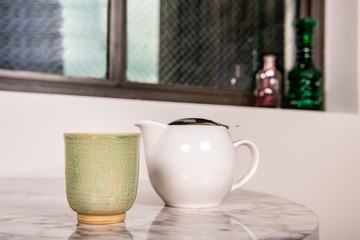 Close up of a tea cup and tea pot on a table in a modern home, by a window with a city view.