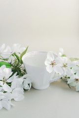 Obraz na płótnie Canvas White porcelain cup with green tea and a fresh white pear tree flower in a lush bouquet of blooming white flowers and green bright leaves on a twig on a light table. vertical image