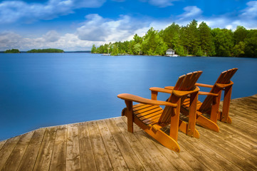 Two Adirondack chairs sitting on a wooden dock facing a blue calm lake. Across the water is a white...