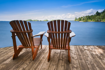 Two Adirondack chairs sitting on a wooden dock facing a blue calm lake. Across the water is a green cabin nestled among green trees. 