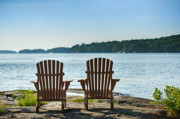 Two Adirondack chairs sit on a rock formation facing the waters of a lake during a sunny summer day in Muskoka, Ontario Canada.
