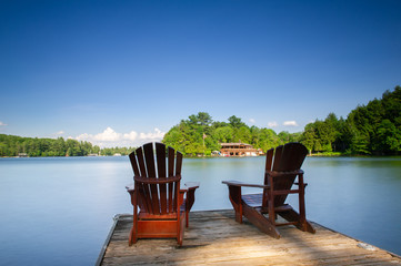 Fototapeta premium Two Adirondack chairs sit on a wooden dock facing a calm lake. The blue water is reflecting the sky clouds. In the background there's a brown cottage nestled between trees. 