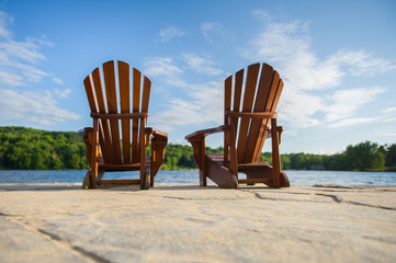 Two Adirondack chairs sit on a stone made pier, facing smooth waters of a lake in Muskoka, Ontario...