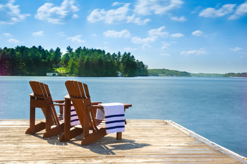 Two Muskoka chairs sitting on a wood dock facing a calm lake. Across the water is a white cottage...