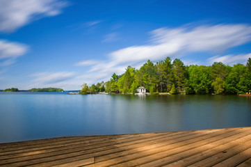 Wood dock in Muskoka facing a lake during a sunny summer day. Across the calm water there's a white...
