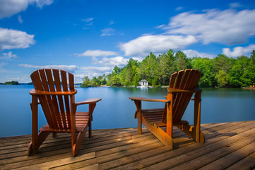 Two Muskoka chairs sitting on a wood dock facing a calm lake. Across the water is a white cottage nestled among green trees. There is a boat dock on the water in front of the cottage. - obrazy, fototapety, plakaty