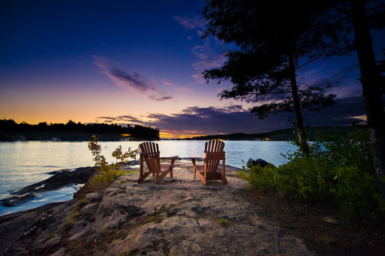 Two Muskoka chairs sitting on a rock formation facing a calm lake at dusk in cottage country. 