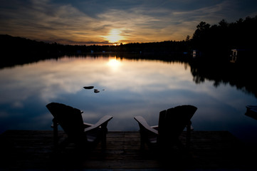 Two  chairs sitting on a wood dock facing a calm lake at sunset. Across the water are cottages...