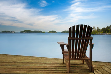 Muskoka chair facing a calm lake on a wooden dock. Across the blue water there's a white cottage.