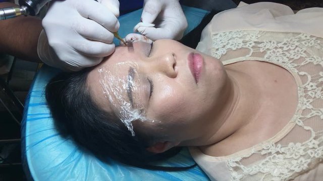 Asia woman tattoo eyebrow by using the electric tattoo machine by beautician professional in beauty salon.