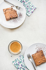 Top view of a cup of coffee and two dessert plates with brownie cake over white rustic background.