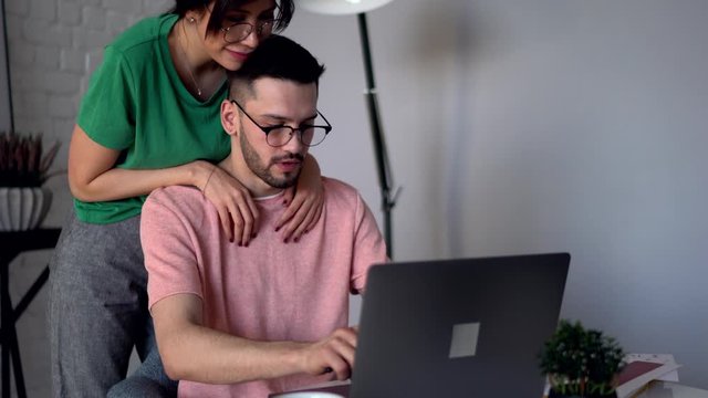 Young woman consoling her husband which working remotely at modern laptop computer in apartment.Lovely girlfriend with tattoo on hand supporting boyfriend sitting at netbook at home interior
