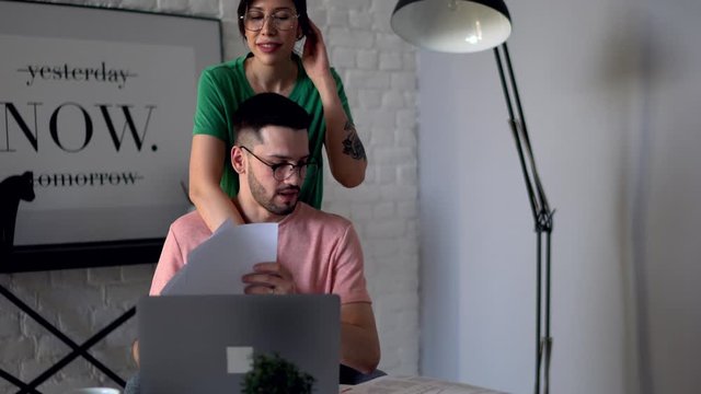 Young bearded man checking financial paper reports working at laptop at home while lovely wife support him.Girlfriend caring boyfriend which sitting at digital netbook in modern apartment 
