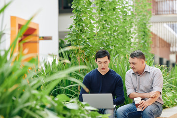 Young Asian entrepreneur showing his project on laptop computer to mentor or mature corporate leader