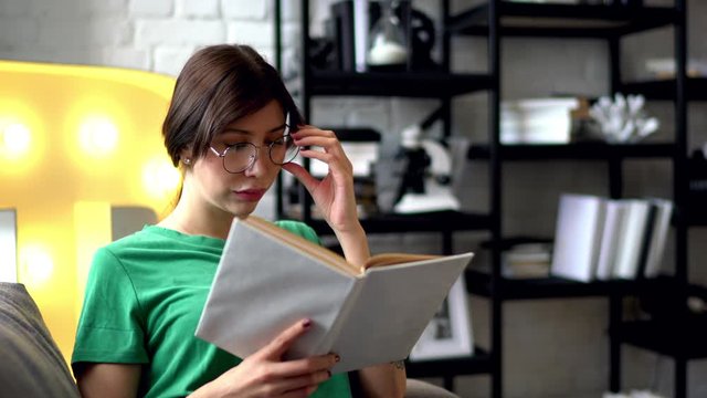 Smart young woman in eyeglasses reading literature book sitting on comfortable couch enjoying recreation time at home interior.Hipster gir resting in free time and learning information from textbook  