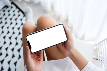 Top view mockup image of woman holding mobile phone with blank desktop white screen while sitting in bedroom with feeling relaxed
