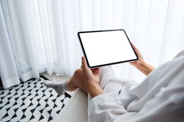 Mockup image of a woman holding black tablet pc with blank white desktop screen while sitting in bedroom with feeling relaxed in the morning