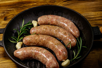 Raw sausages ready for preparation with rosemary, garlic and spices in cast iron grill frying pan on wooden table