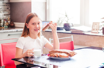 A girl sits in the kitchen in a white T-shirt, drinks milk from a cup and eats homemade bread