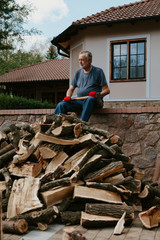 Elderly man in red gloves sits over pile of firewood