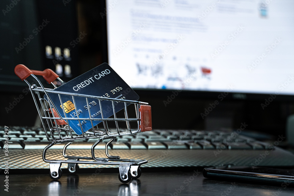 Poster close up Shallow focus Credit Card in mini shopping card with Computer keyboard and E-commerce website on monitor background. Background for E-commerce and online shopping concept.