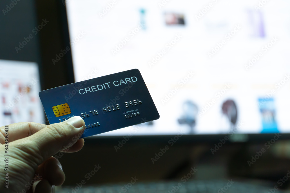 Poster close up Shallow focus hand hold Credit Card with Computer keyboard and E-commerce website on monitor background. Background for E-commerce and online shopping concept.