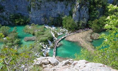 Plitvice National Park in Croatia, beautiful landscape with waterfalls, long line of tourists walking on special wooden footbridge over emerald color lakes.