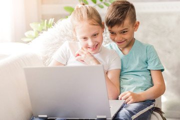 Happy brother and sister sitting on sofa at home in the living room and using laptop together.