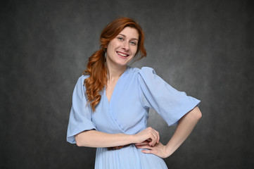 The model poses with different emotions on a gray background in the studio. Portrait of a young caucasian woman with long red hair in a blue dress.
