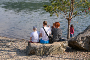 People sit on a stone in summer. View from back. Mom, dad, kid. Concept of a family vacation on shore of a pond.