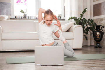 A little girl of school age does sports exercises on a gymnastic carpet on the floor. Nearby is a laptop, there is a video with gymnastic exercises.