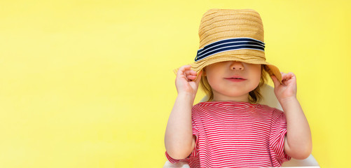 Little toddler girl hides her face with a straw hat. The child pulled a hat on his head on a yellow background. Copy space and place for text