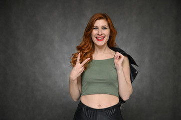 Pretty model actress posing with different emotions on a gray background in the studio. Portrait of a young Caucasian woman with long red hair in a green T-shirt and with a black jacket in her hands.
