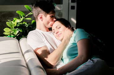 Lovers relax on a cozy sofa in the living room, a woman lies on the man s shoulder, a man and a woman smile and spend a lazy weekend. The room has bright sunlight and hard shadows.