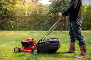 A worker cutting grass in green yard at sunset. Man with electric lawnmower, lawn mowing. Gardener trimming a garden