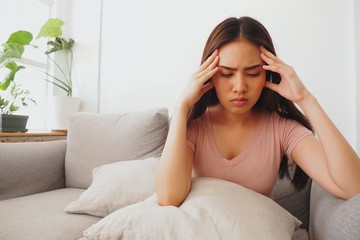Young Asian woman sitting on sofa at home using her hands touching her head feeling tired and stressed about her work and health. Having fever and headache due to insufficient sleep and health problem