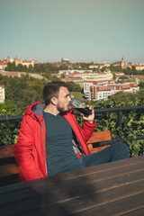 Fototapeta premium Man drinking beer. Handsome young hipster man drinking beer while sitting at the bar outdoors with panoramic view of Prague